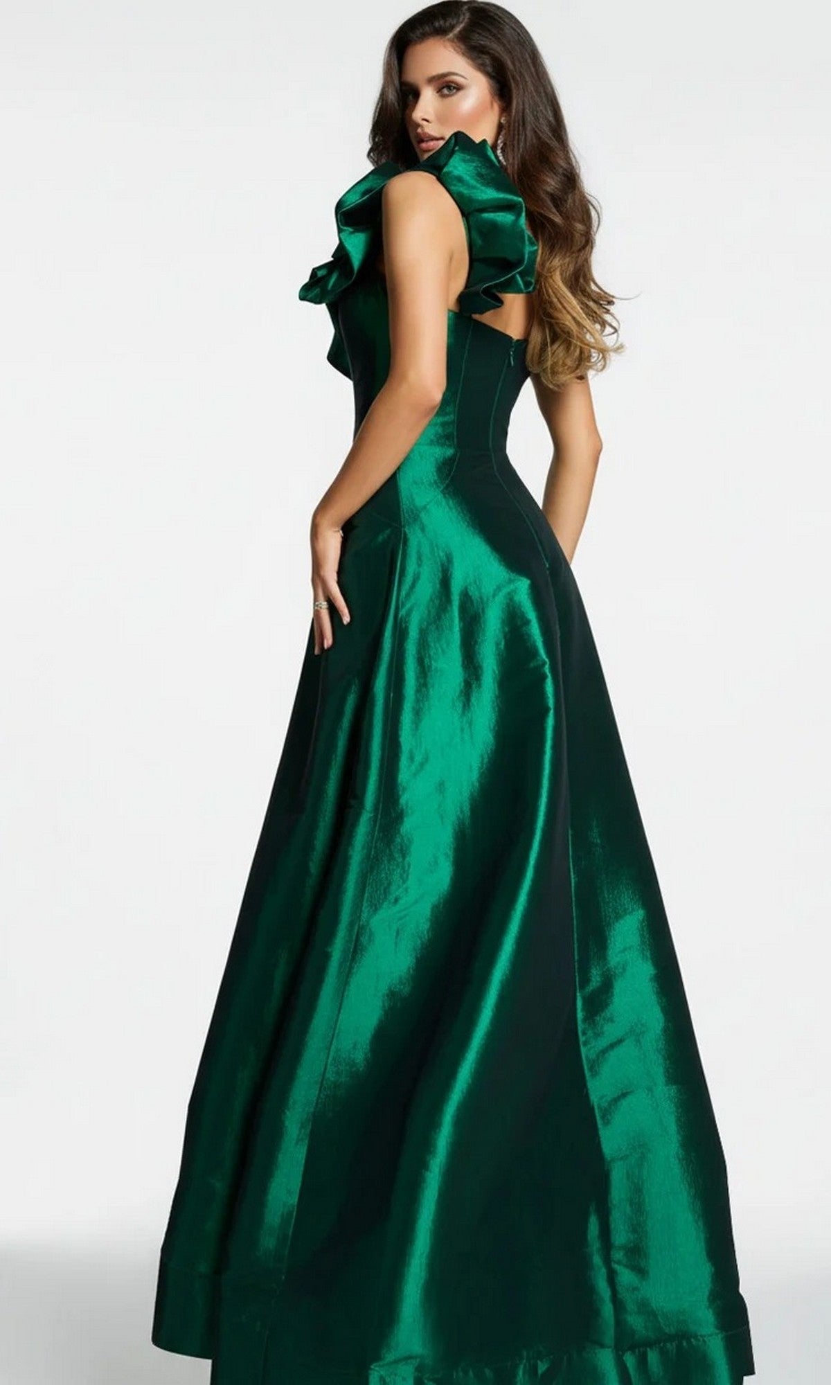 A woman wearing the Long Formal Dress: Jovani 43048, a shiny emerald green satin evening gown featuring ruffle shoulder detail, stands against a plain background and looks over her left shoulder.